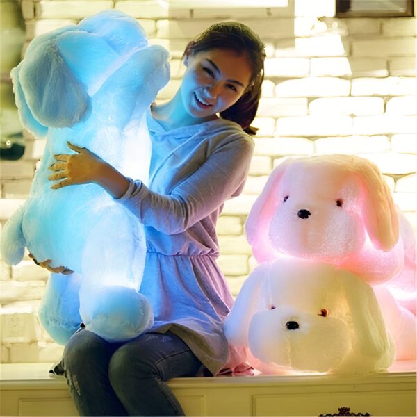 peluche-chien-lumineux-bleu-jouet-en-peluche-lumineux-3a-2 Une jeune femme souriante tenant un grand chien en peluche bleu lumineux et deux autres chiens en peluche rose et blanc sur un banc.