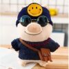 peluche-cochon-smiley-bonnet-et-lunettes-de-soleil-2a-2 Une peluche de cochon portant un bonnet bleu avec un smiley jaune, des lunettes de soleil rondes et un pull bleu avec un sac en bandoulière marron.