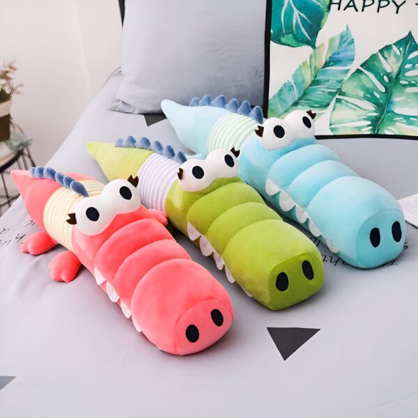 Trois peluches crocodiles en forme de coussin, de couleur rose, verte et bleue avec des rayures blanches et des yeux noirs.