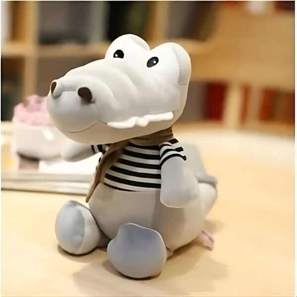 Peluche de crocodile gris avec un foulard brun et un t-shirt rayé noir et blanc assis sur une table en bois.