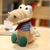 peluche-crocodile-bebe-marin-jouet-doux-enfant-5a Une peluche de crocodile beige avec des yeux noirs et un foulard rouge, portant un t-shirt à rayures bleues et blanches, assis sur un bureau en bois.