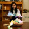peluche-dauphin-lumineuse-lampe-de-chevet-et-compagnon-de-sommeil-8a Une jeune femme souriante assise sur le sol avec quatre peluches dauphins lumineux. Les dauphins sont de différentes couleurs, et ils brillent de différentes couleurs.