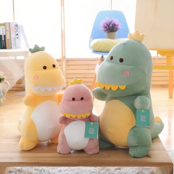 peluche-dinosaure-amusant-jouet-doux-pour-enfant-2a-2 Trois dinosaures en peluche, un jaune, un rose et un vert, sont assis sur un plancher en bois. Les trois peluches ont un sourire amical et des yeux noirs.
