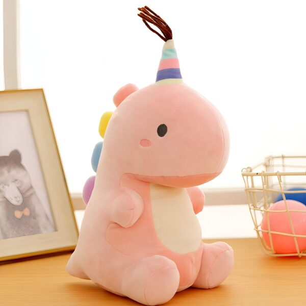 Une peluche rose en forme de dinosaure avec un chapeau arc-en-ciel sur la tête. Elle est assise sur une table en bois.