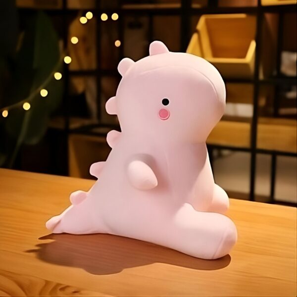peluche-dinosaure-rose-jouet-doux-et-calin-pour-enfants-4a Peluche rose en forme de dinosaure avec des yeux noirs et un nez rose, sur un fond en bois.