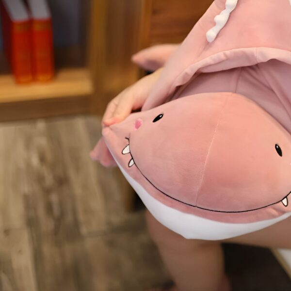 peluche-dinosaure-rose-magique-2a-2 Un gros coussin en peluche rose en forme de requin avec de grandes dents blanches et des yeux noirs.