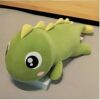 Peluche de dinosaure vert avec de grands yeux noirs et des joues roses. Il est allongé sur le dos.