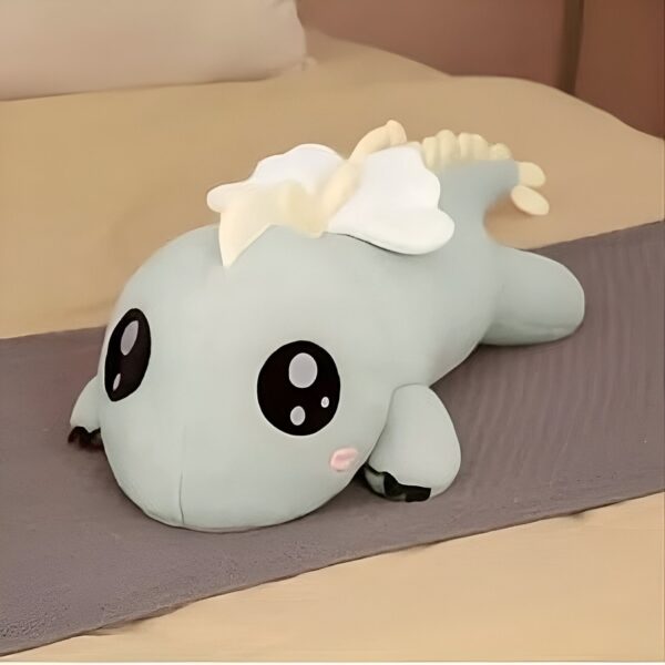 Peluche de dragon vert avec de grands yeux noirs, allongé sur un lit gris.