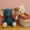 Un éléphant en peluche bleu et un chiot en peluche blanc et brun assis sur un fond rose. Le chiot est assis dans un panier en osier avec une doublure rouge et blanche à carreaux.