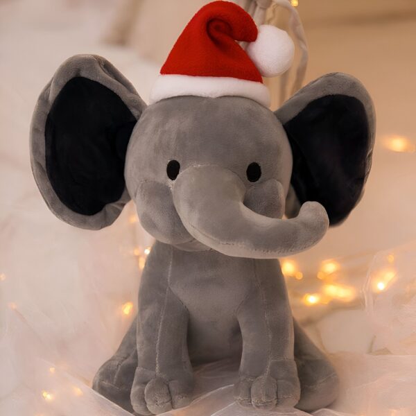 Un éléphant en peluche gris portant un chapeau de Père Noël, assis sur un tissu blanc avec des lumières scintillantes.