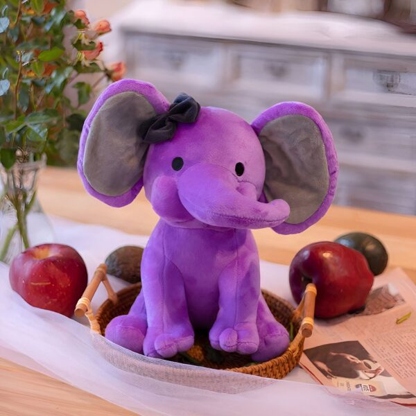 Un éléphant en peluche violet avec un nœud noir sur la tête, assis dans un panier en osier sur une table en bois.
