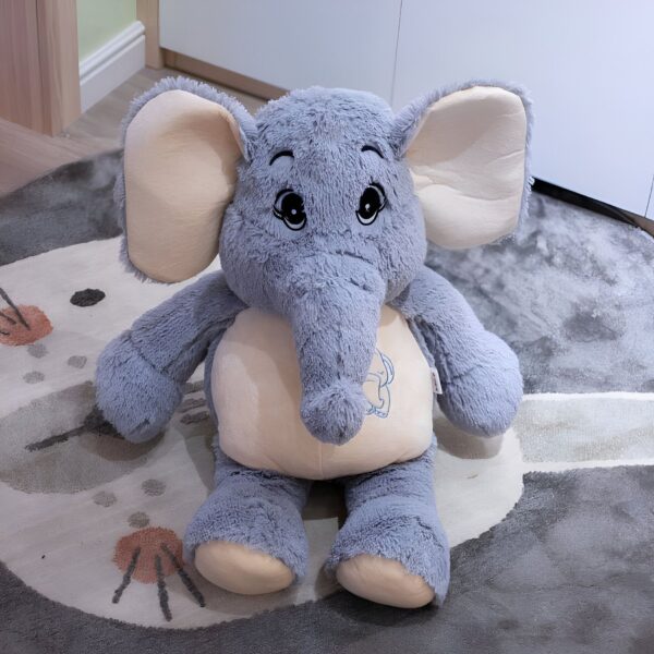 Une grande peluche d'éléphant gris avec de grandes oreilles et des yeux noirs. Il est assis sur un tapis gris.