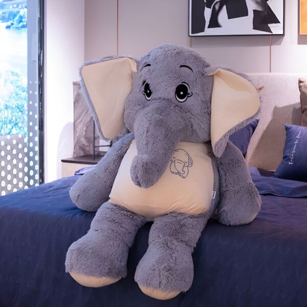 Une grande peluche d'éléphant gris assis sur un lit avec un petit éléphant brodé sur son ventre.