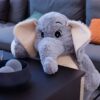 Un éléphant en peluche gris avec de grandes oreilles beige repose sur un canapé et s'appuie sur une table.