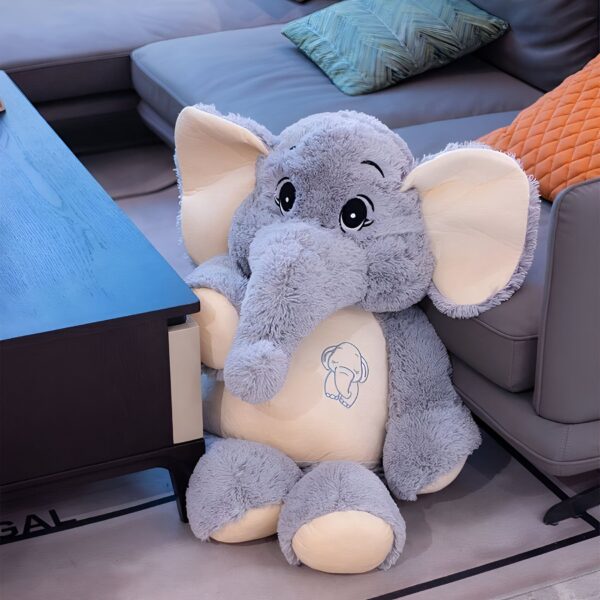 Grande peluche d'éléphant gris assis sur un canapé. L'éléphant a de grands yeux noirs, une petite trompe et un dessin d'éléphant sur la poitrine.