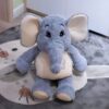 Une peluche d'éléphant gris avec de grands yeux noirs et de grandes oreilles. L'éléphant est assis sur un tapis gris clair avec des motifs.