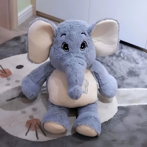 Une peluche d'éléphant gris avec de grands yeux noirs et de grandes oreilles. L'éléphant est assis sur un tapis gris clair avec des motifs.