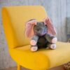 Un éléphant en peluche gris avec des oreilles roses et un nœud violet assis sur un fauteuil jaune.