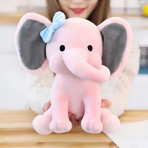 Une peluche rose en forme d'éléphant avec un nœud bleu sur la tête, tenue par une personne.