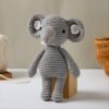 Un adorable éléphant en crochet gris avec de grandes oreilles et un petit tronc. Il est debout sur un bloc de bois.