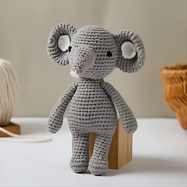 Un adorable éléphant en crochet gris avec de grandes oreilles et un petit tronc. Il est debout sur un bloc de bois.