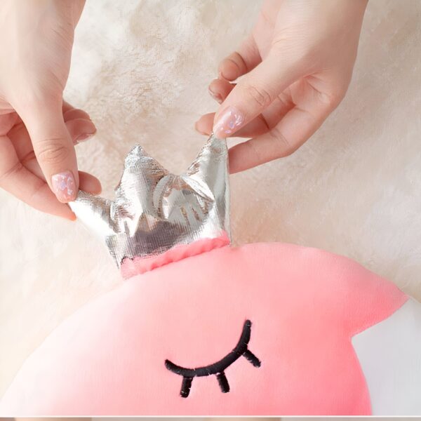 Un oreiller rose en forme de dauphin avec une couronne argentée sur la tête, tenu par deux mains.