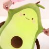 Un gros coussin en forme d'avocat souriant avec des yeux et des joues rouges. Il est en peluche verte avec un noyau brun.