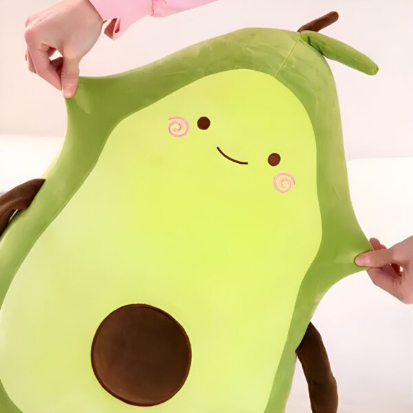 Un gros coussin en forme d'avocat souriant avec des yeux et des joues rouges. Il est en peluche verte avec un noyau brun.