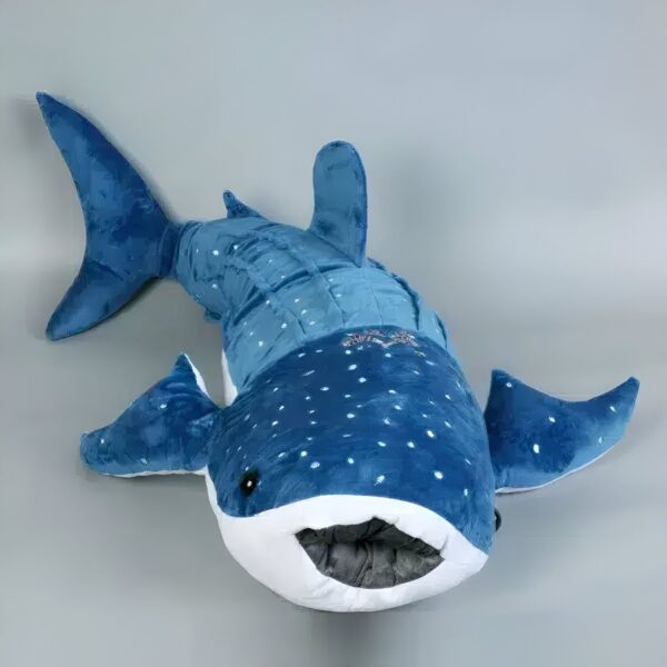 Une grande peluche bleue en forme de requin-baleine avec des points blancs. Le requin a une bouche ouverte et des nageoires pectorales.