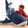 Un jeune garçon en rouge câline une grande peluche en forme de requin baleine bleu à pois blancs.