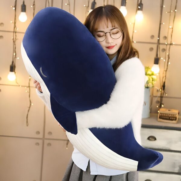 Une jeune femme en lunettes serre dans ses bras une grande peluche en forme de baleine bleue.