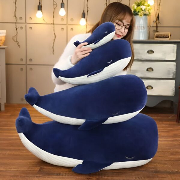 Trois baleines en peluche bleues empilées l'une sur l'autre. La plus grande baleine est sur le sol et la plus petite est sur les genoux d'une femme.