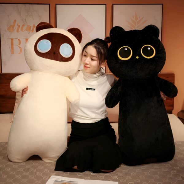 Une jeune femme est assise sur un lit et tient deux grandes peluches en forme de chat, l'une blanche et l'autre noire.