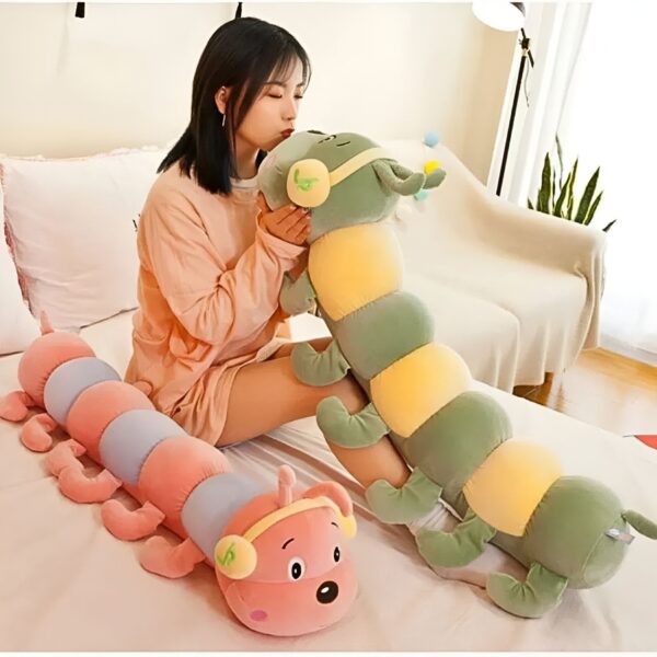 Une jeune femme souriante tenant deux gros peluches en forme de chenilles. La chenille verte est plus grande et la chenille rose est plus petite. Les deux peluches ont des écouteurs.