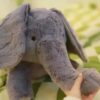 Un éléphant en peluche gris, avec de grandes oreilles, se trouve sur un tissu vert et jaune. La main d'une personne est visible en bas à droite de l'image, tenant l'éléphant par la patte.
