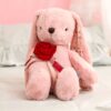 Un lapin rose en peluche tenant une rose rouge dans ses pattes. La peluche est assise sur un lit blanc.
