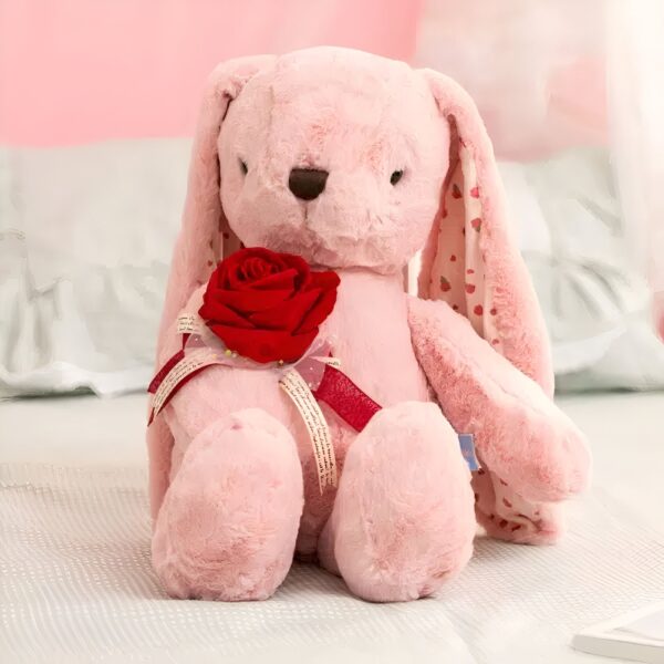 Un lapin rose en peluche tenant une rose rouge dans ses pattes. La peluche est assise sur un lit blanc.