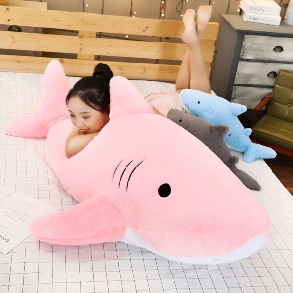 Une jeune femme souriante allongée sur un lit avec une grande peluche de requin rose. Elle a aussi deux autres peluches de requins, une bleue et une grise, sur le lit.