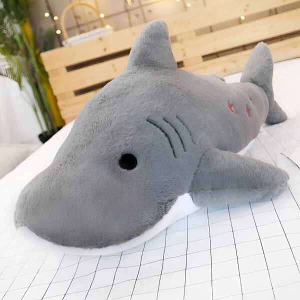 Une peluche de requin gris avec des yeux noirs et une bouche blanche sur un lit blanc.