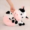 Une peluche de vache noire et blanche avec un visage rose et des yeux noirs. La vache est couchée sur le côté, une main humaine la serre contre elle.
