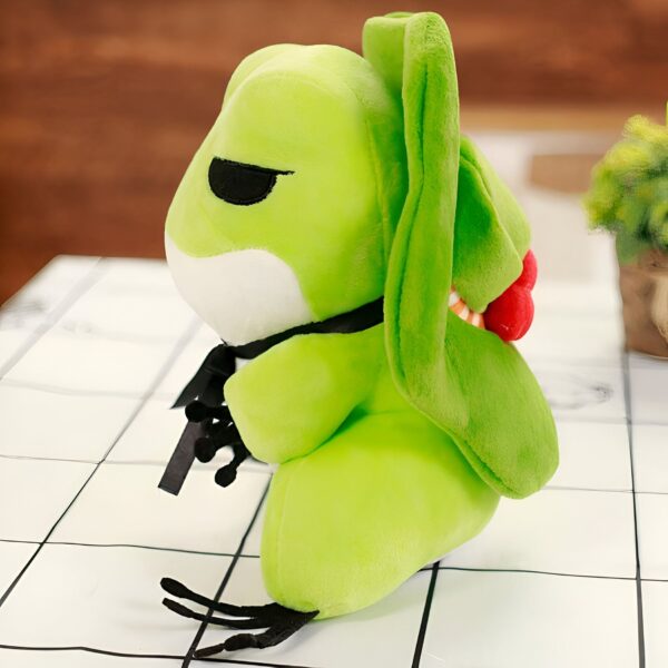Une grenouille en peluche verte avec un nœud noir et une bouche rouge. Elle a de grands yeux noirs et des pattes noires.