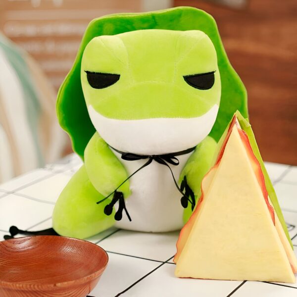 Une grenouille en peluche verte avec un ruban noir autour du cou, assise à côté d'un sandwich triangulaire.