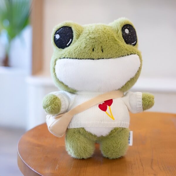 Une grenouille en peluche verte portant un pull blanc avec un motif de cœur rouge et une bandoulière beige.