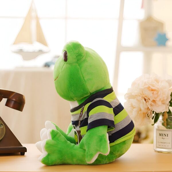 Une grenouille en peluche verte portant un t-shirt rayé bleu et vert assis sur une table en bois.
