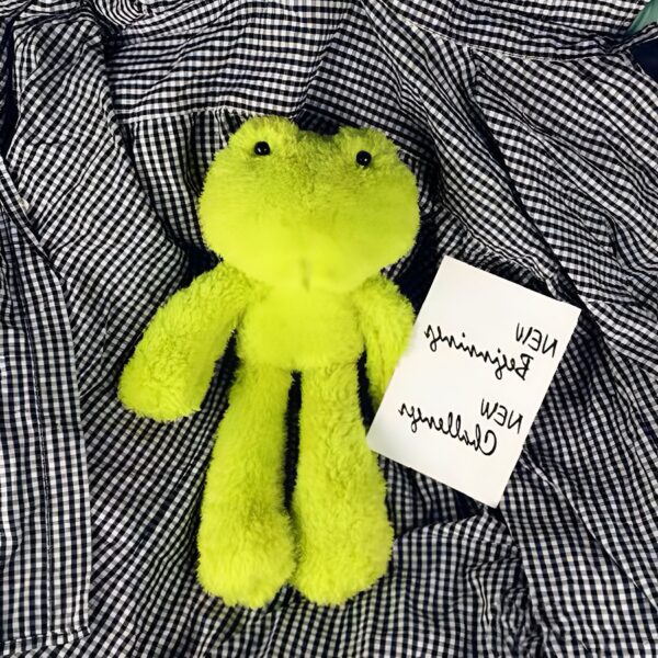 peluche-grenouille-verte-jouet-doux-pour-enfant-ideal-contre-la-peur-du-sommeil-2a-2 Une grenouille en peluche verte sur un tissu à carreaux noirs et blancs.