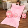 Une couverture rose en peluche avec trois hiboux en peluche. Les hiboux sont de différentes tailles et sont tous assis sur la couverture. La couverture est posée sur un canapé en bois.