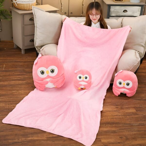 Une couverture rose en peluche avec trois hiboux en peluche. Les hiboux sont de différentes tailles et sont tous assis sur la couverture. La couverture est posée sur un canapé en bois.