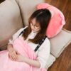 Une jeune femme portant des lunettes se repose sur un canapé confortable avec un oreiller en forme de chat rose et une couverture rose.