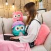 Une jeune femme assise sur un canapé avec un ordinateur portable sur ses genoux. Elle porte une robe blanche à carreaux noirs. Elle est entourée de trois peluches en forme de hibou.