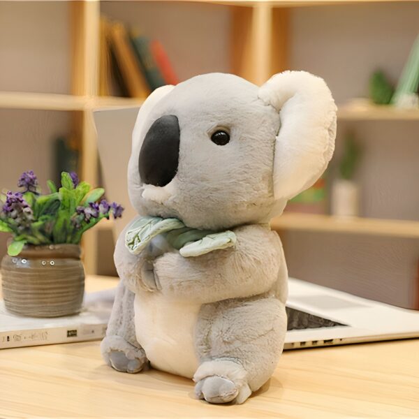 Une peluche koala grise et blanche assise sur une table en bois, tenant une feuille verte dans ses pattes.